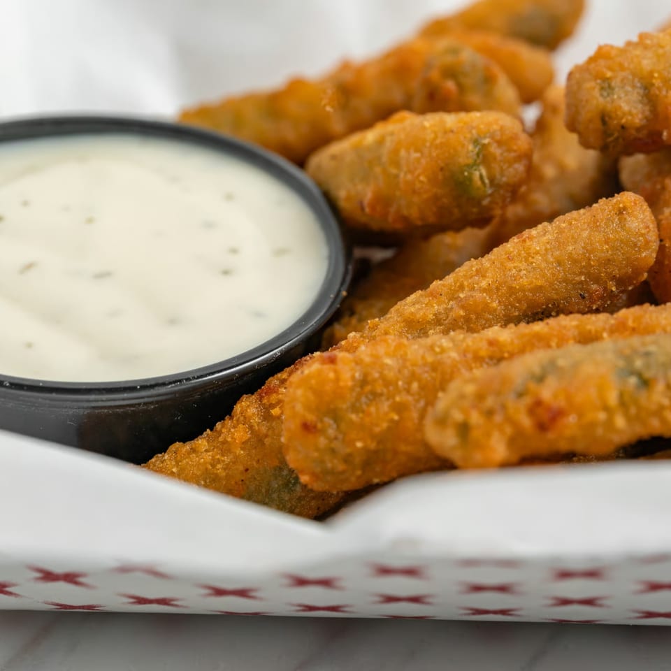 Crispy fried pickle spears in a paper container with a side of creamy ranch dipping sauce.