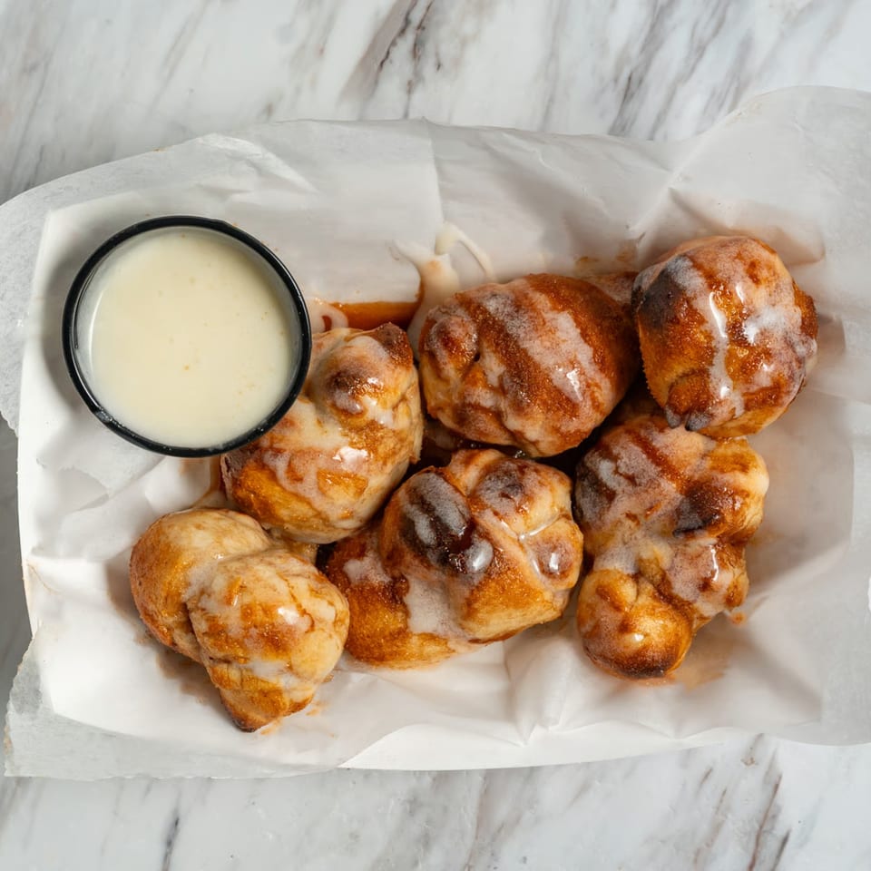 Seven glazed cinnamon sugar donut holes in a paper-lined basket with a side of dipping sauce.