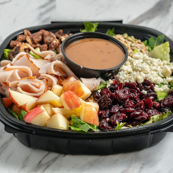 A large salad with turkey, apples, cranberries, blue cheese, nuts, and dressing in a black container.