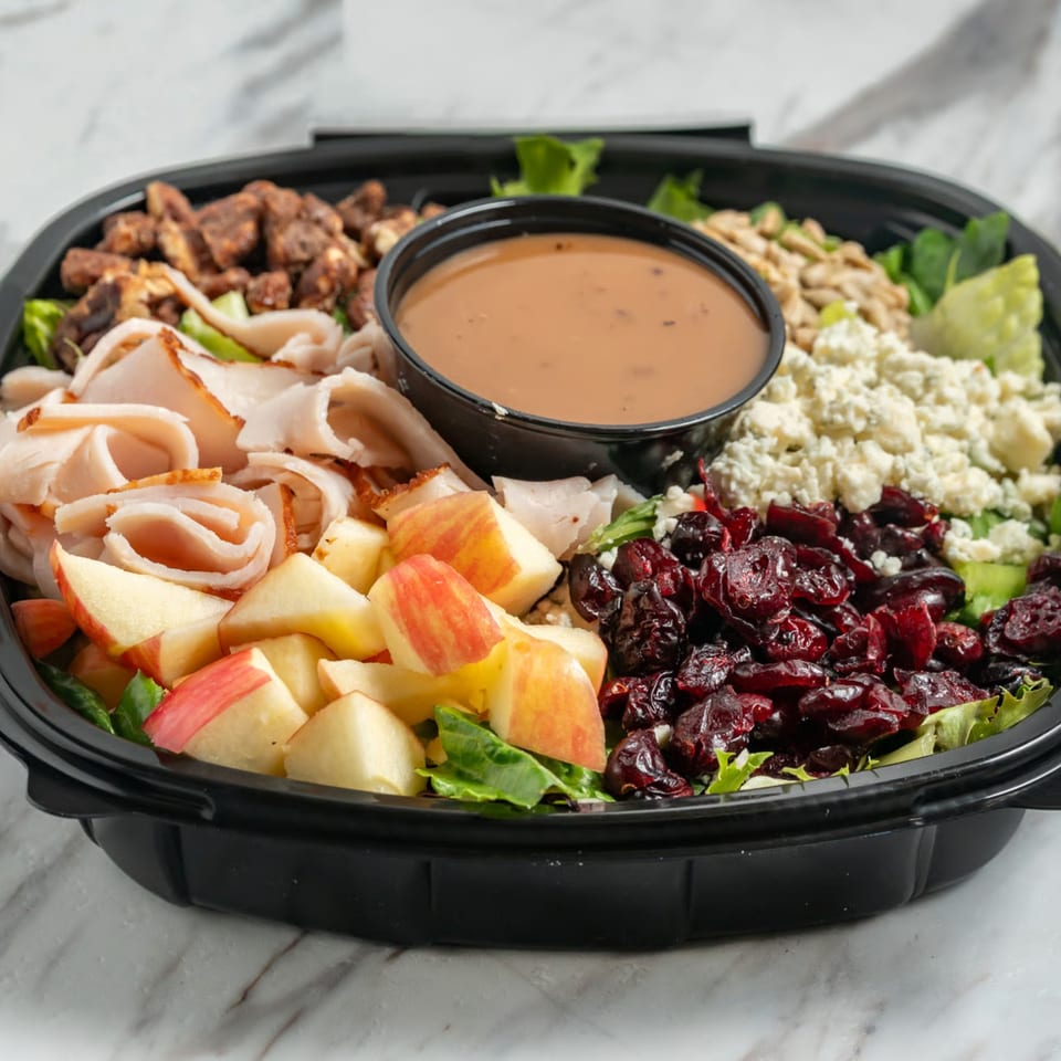 A large salad with turkey, apples, cranberries, blue cheese, nuts, and dressing in a black container.