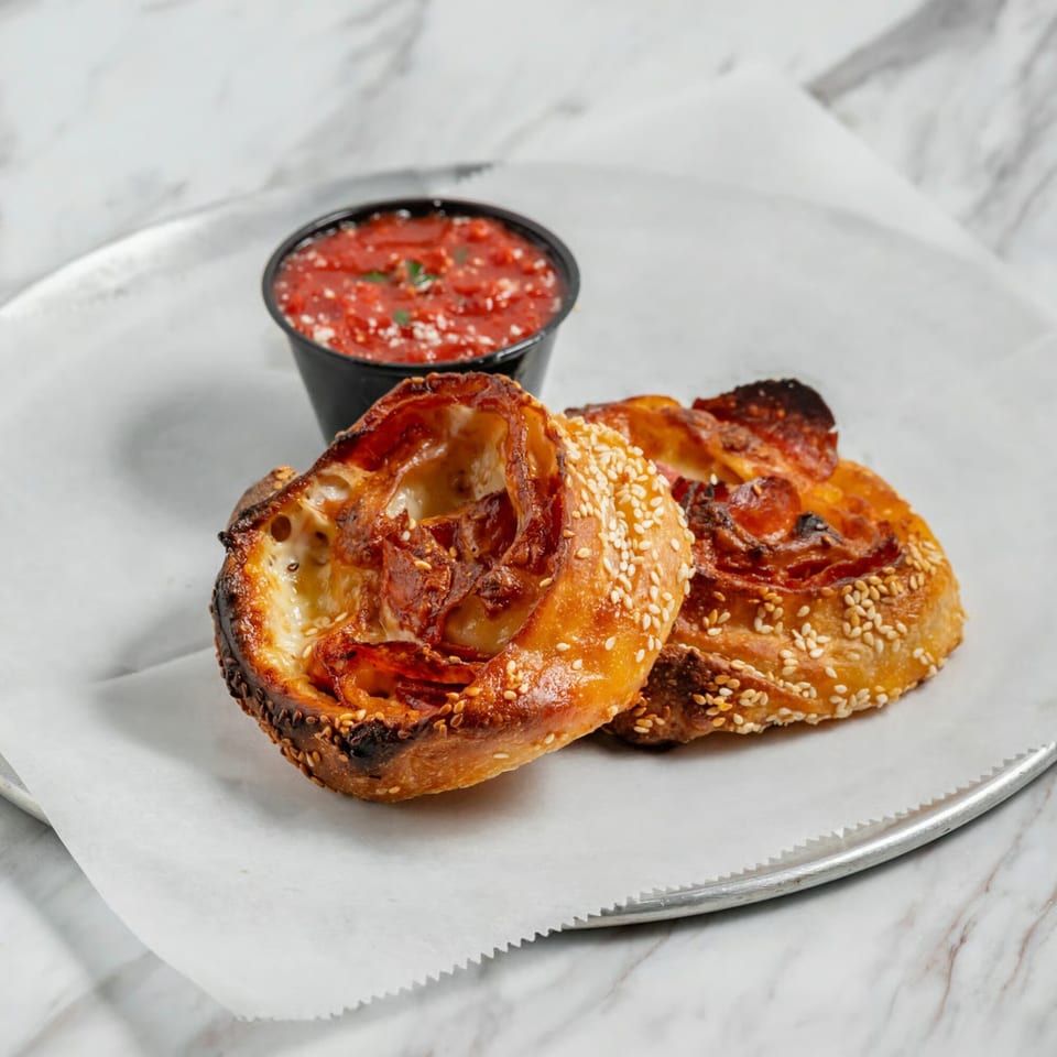 Two golden-brown pizza rolls with sesame seeds and pepperoni, served with a side of marinara sauce.